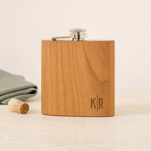May include: A rectangular wooden flask with a light brown grain pattern and a silver-toned metal screw cap. The front surface features the engraved letters K and R separated by a vertical line. The flask has a smooth finish and rounded edges.