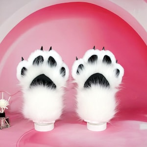 Peut inclure: Une paire de gants en fausse fourrure blanche en forme de pattes avec des coussinets noirs et des griffes noires. Les gants sont conçus pour ressembler à des pattes d'animaux. Sur fond rose.
