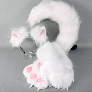 Op de afbeelding: Een set witte imitatiebont dierenaccessoires bestaande uit een haarband met twee puntige oren met roze binnendetails, een paar pluche poothandschoenen met roze voetzooltjes en witte klauwen, en een lange, gebogen, pluizige witte dierenstaart.