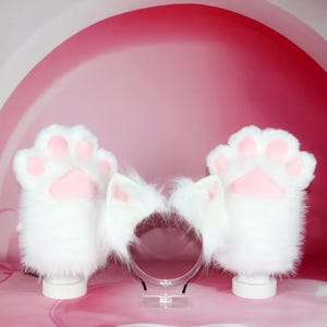 May include: A set of white, fluffy cat paw gloves and a matching headband with cat ears. The gloves have pink paw pads. The items are displayed on white stands against a pink background.