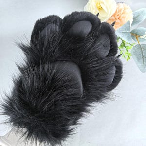 May include: A black faux fur paw with four padded pads and sharp claws. The furry paw is a costume accessory, likely for cosplay or themed events. The paw is shown against a soft, neutral background with floral accents.