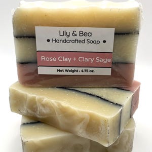 May include: Stacked bars of handcrafted soap with a label that reads "Lily & Bea Handcrafted Soap". The soap has layers of cream, pink, and black. The label also says "Rose Clay + Clary Sage" and "Net Weight: 4.75 oz."