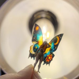 May include: A miniature figurine of a nude woman with butterfly wings. The wings are a vibrant mix of blue, yellow, and orange, with celestial and sun designs. The figure is held against a bright light.