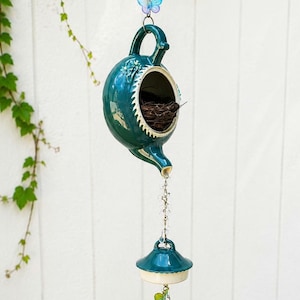 May include: A teal ceramic teapot birdhouse hanging from a chain with a blue butterfly accent. The teapot features a circular opening containing a small nest, a spout with a crystal strand, and a matching lid hanging below with silver bells.