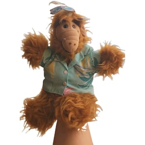May include: A brown, furry plush puppet with a long snout and dark eyes, wearing a light blue short-sleeved button-down shirt featuring a tropical pattern with yellow and green shapes, and a pair of blue-tinted sunglasses resting on its head.