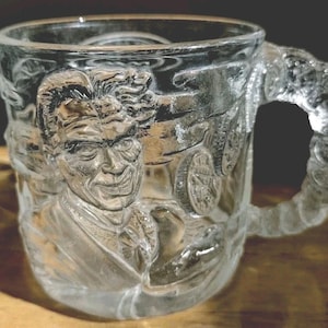 May include: Clear glass mug with a detailed embossed design featuring a smiling face, a car, and other elements. The handle is also embossed with a decorative pattern. The mug is transparent, allowing the contents to be visible.
