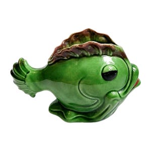 May include: A ceramic fish-shaped planter in glossy green with brown accents. The fish has a wide open mouth and a large eye. The planter is designed to hold small plants or flowers, adding a whimsical touch to any space.