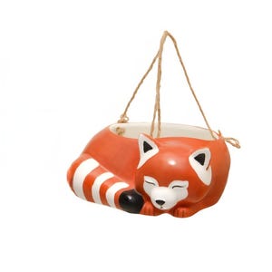 May include: A ceramic hanging planter shaped like a sleeping red panda. The planter features a vibrant orange body, a white face with black accents, and a striped tail. It is suspended by thin, natural-colored jute twine attached to the rim.