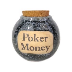 May include: A round, speckled black ceramic jar with a cork stopper. The jar has a tan, rectangular label with the words "Poker Money" in brown, stylized lettering. The jar is designed to hold poker winnings.