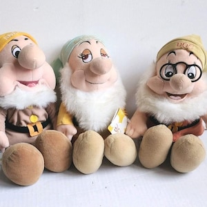 May include: Three plush dwarf dolls from Snow White. Each doll has a round, tan body, a white beard, and a hat. One doll wears glasses. The dolls are in various colors, including yellow, green, and brown.