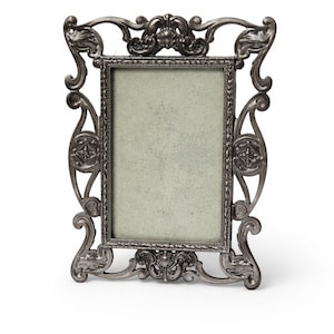 May include: Ornate silver-colored picture frame with a rectangular opening. The frame features elaborate scrollwork and decorative flourishes. The frame is designed to hold a photograph or artwork. The frame is made of metal.
