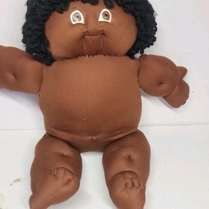 May include: A brown fabric doll with black curly hair. The doll has a round face with large, light-colored eyes and a small smile. The doll's arms and legs are also brown, and it has a simple, stitched design.