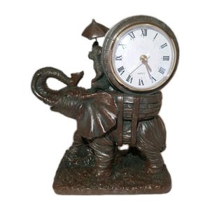 May include: A bronze-colored elephant figurine with a clock face. The clock has Roman numerals and gold-colored hands. A small figure with an umbrella sits atop the elephant. The elephant is standing on a textured base.
