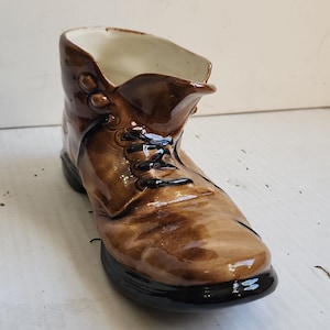 May include: A brown ceramic boot-shaped planter or decorative vessel with a glossy finish. The boot features dark brown laces, a smooth toe area, and a solid black sole. The interior of the boot is glazed in a clean, off-white color.