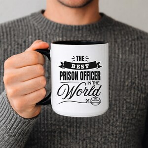 Puede incluir: Taza de cerámica blanca con asa y borde negros. La taza presenta el texto "THE BEST PRISON OFFICER IN THE World" en escritura negra y un gráfico de insignia.