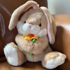 May include: A plush bunny rabbit with long ears, holding a yellow and orange flower. The bunny has a light brown fur with white accents on its face and paws. The inside of the ears is pink.