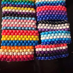May include: Several beaded bracelets in various colors, including rainbow, pink, blue, and white. The bracelets are made of small, round beads strung together, and are arranged in stacks. The bracelets appear to be handmade and are likely intended for fashion or self-expression.