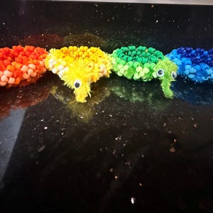 May include: A row of colorful, handmade worm toys in a rainbow of colors: red, orange, yellow, green, blue, and purple. Each worm has googly eyes and a fuzzy head. These whimsical toys are likely made from pipe cleaners and beads.