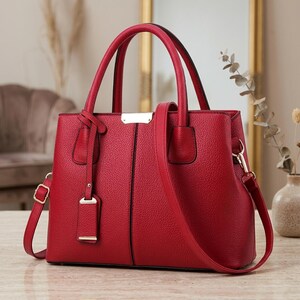 May include: A bright red handbag with a textured, pebbled surface, featuring two top handles, a detachable shoulder strap, and a central vertical black seam. The bag includes a gold-toned metal accent plate at the top and a matching hanging tag.