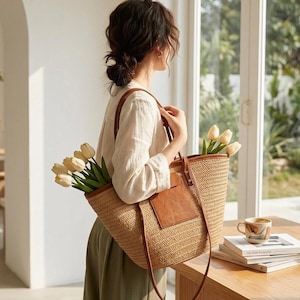 May include: A large woven straw tote bag with brown leather handles and a matching leather patch on the front. The bag is filled with a bouquet of cream-coloured tulips, featuring a textured natural fibre construction and long shoulder straps.
