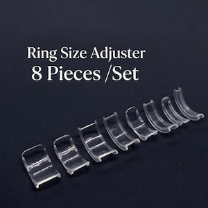 Puede incluir: Un conjunto de ocho ajustadores de tamaño de anillo de plástico transparente. Los ajustadores están dispuestos en diagonal sobre un fondo oscuro. El texto en la parte superior dice "Ring Size Adjuster 8 Pieces / Set".