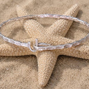 May include: A silver-colored hammered metal bracelet with a hook clasp rests on a beige starfish, set against a sandy background. The bracelet has a textured surface and a simple, elegant design, perfect for beach-themed jewelry.