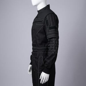 K&ouml;nnte beinhalten: Schwarzer Overall mit wei&szlig;en Steppn&auml;hten. Er hat einen hohen Kragen, gepolsterte Schultern und geometrische Muster an den &Auml;rmeln. Der Anzug hat mehrere Taschen und einen G&uuml;rtel. Das Design ist modern und strukturiert.