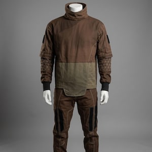 May include: Brown two-piece outfit with a high collar. The top has a brown body with olive green accents and geometric patterned sleeves. The pants have black accents. The outfit is displayed on a mannequin.