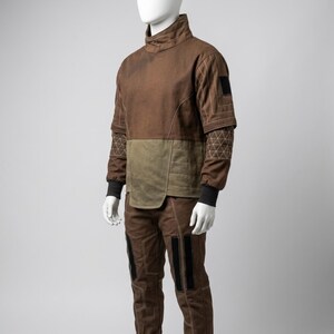 May include: A brown and olive green two-piece outfit displayed on a mannequin. The top has a high collar and quilted accents, with black cuffs. The pants have black panels on the legs. The outfit is designed with visible stitching.