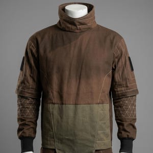 May include: A brown jacket with a high collar and geometric stitching details. The jacket features a two-tone design with an olive green lower section and black cuffs. The sleeves have a layered design with a geometric pattern on the lower sleeves.