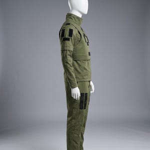 May include: Olive green military-style uniform displayed on a mannequin. The outfit includes a high-collared jacket, a vest with black rectangular patches, long sleeves, and matching pants. The pants also feature black patches. The uniform is made of a durable fabric.