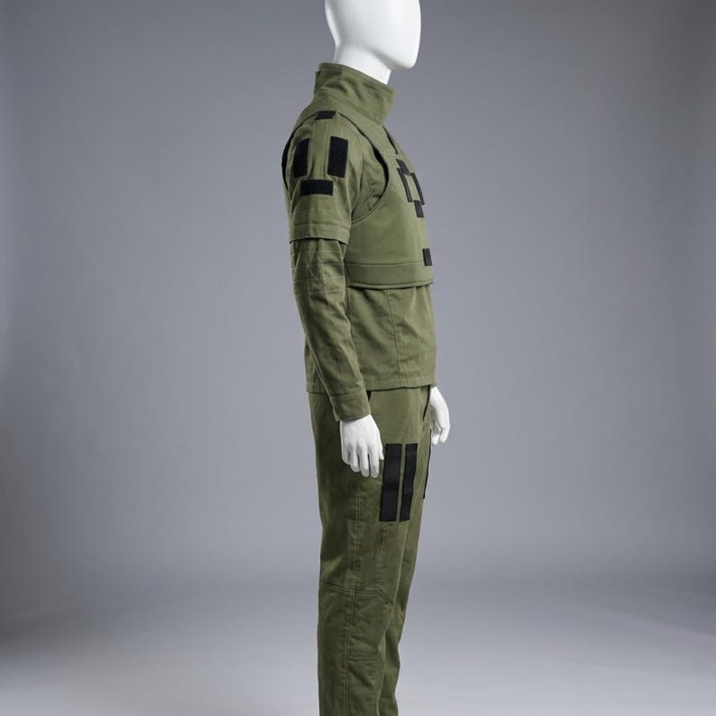 May include: Olive green military-style uniform displayed on a mannequin. The outfit includes a high-collared jacket, a vest with black rectangular patches, long sleeves, and matching pants. The pants also feature black patches. The uniform is made of a durable fabric.