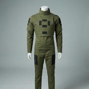 May include: Olive green tactical uniform displayed on a mannequin. The outfit includes a high-neck shirt, a vest with black square patches, and matching pants. The long-sleeved shirt has shoulder pads, and the pants have knee patches. The uniform is designed for functionality.