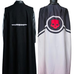 Könnte beinhalten: Zwei Umhänge, einer schwarz mit weißem Emblem und der andere mit rotem Totenkopf-Design. Der schwarze Umhang hat Lederärmel und ein weißes Emblem mit Totenkopf und Flügeln. Der zweite Umhang hat einen roten Totenkopf in einem weißen Kreis auf schwarz-weißem Hintergrund.