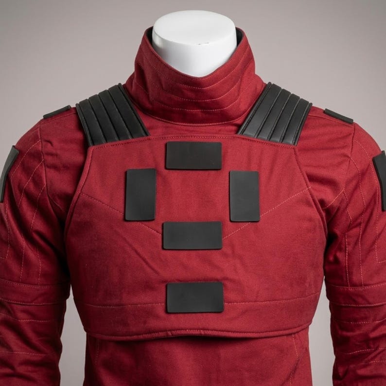 May include: A red jacket with a high, ribbed collar and black accents. The jacket features black rectangular patches on the chest and shoulders. The garment is displayed on a white mannequin head and shoulders.