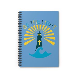 May include: A spiral-bound notebook with a light blue cover featuring a lighthouse design. The lighthouse is black with a yellow cross and is set against a backdrop of yellow rays and a blue wave. The text "BE THE LIGHT" is above the lighthouse.