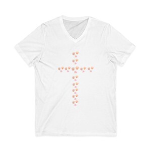 May include: White V-neck t-shirt featuring a cross design made of seashells and starfish. The cross is centered on the chest and is composed of various sizes of light orange seashells and pink starfish.