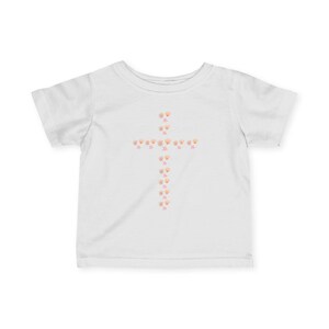 May include: White t-shirt featuring a cross design made of peach-colored seashell graphics and small pink star accents. The short-sleeved shirt is made of a soft material, suitable for children.