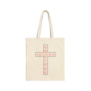 May include: A cream-coloured tote bag with a black outline of a cross filled with small red hearts. The bag has two long handles for carrying over the shoulder. The design is centred on the front of the bag.
