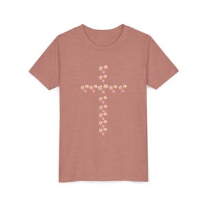 May include: A short-sleeved, heathered pink t-shirt featuring a cross design made of seashells and pink star accents. The cross is centered on the chest of the shirt. The shirt is made of a soft material.