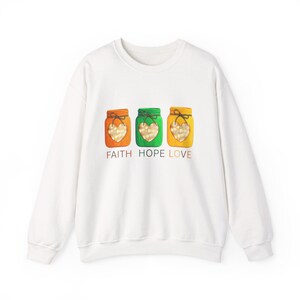 Pode incluir: Uma camisola branca de manga comprida com um gráfico de três frascos coloridos em laranja, verde e amarelo. Cada frasco tem um recorte em forma de coração preenchido com pequenas luzes brancas e o texto FAITH HOPE LOVE impresso abaixo.