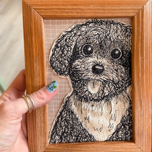 Op de afbeelding: Een ingelijst kunstwerk met een zwart-wit geborduurd hondenportret. De hond heeft grote ogen en een uitgestoken tong. Het kunstwerk is gevat in een lichtbruine houten lijst.