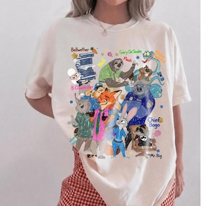 Op de afbeelding: Cr&egrave;mekleurig T-shirt met een kleurrijke afbeelding van verschillende stripfiguren met hun namen. Het ontwerp bevat personages als Bellwether, Gary DeSnake, Flash en Chief Bogo. Het shirt wordt door een persoon gedragen.