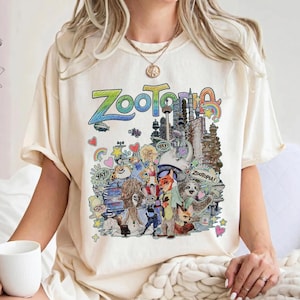 May include: Cream-colored t-shirt featuring a colorful graphic of the word "Zootopia" and various characters from the animated film. The design includes cityscapes, rainbows, and cartoon animals. The shirt is a relaxed fit.