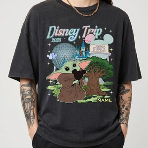 Puede incluir: Camiseta gris oscuro con un gr&aacute;fico de Baby Yoda sosteniendo una cabeza de Mickey Mouse, con el texto "Disney Trip 2026" e im&aacute;genes de parques Disney. La camiseta es de corte holgado.