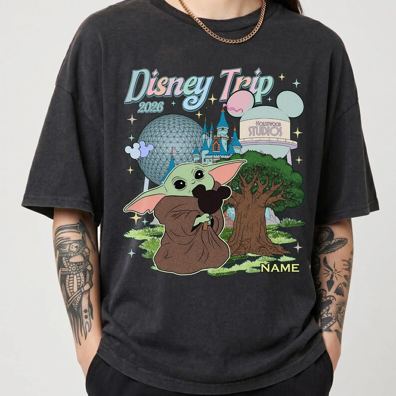 Puede incluir: Camiseta gris oscuro con un gr&aacute;fico de Baby Yoda sosteniendo una cabeza de Mickey Mouse, con el texto "Disney Trip 2026" e im&aacute;genes de parques Disney. La camiseta es de corte holgado.