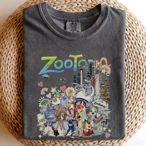 May include: Dark gray Comfort Colors t-shirt featuring a colorful graphic from the movie Zootopia. The design includes the word "Zootopia" in large, stylized letters, along with various characters and elements from the film. The shirt is folded on a woven surface.