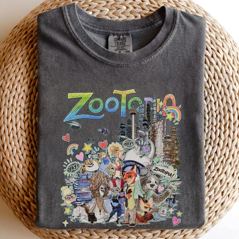 May include: Dark gray Comfort Colors t-shirt featuring a colorful graphic from the movie Zootopia. The design includes the word "Zootopia" in large, stylized letters, along with various characters and elements from the film. The shirt is folded on a woven surface.
