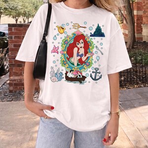 以下が含まれることがあります： 白いTシャツ。装飾的なフレームの中に人魚のカラフルなイラストが描かれており、ボート、錨、泡などの水生要素に囲まれています。デザインには城、フォーク、楽器が含まれています。
