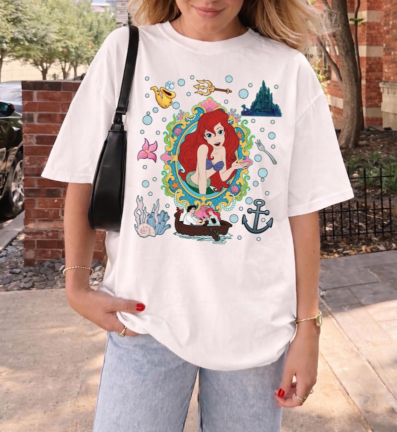 以下が含まれることがあります： 白いTシャツ。装飾的なフレームの中に人魚のカラフルなイラストが描かれており、ボート、錨、泡などの水生要素に囲まれています。デザインには城、フォーク、楽器が含まれています。